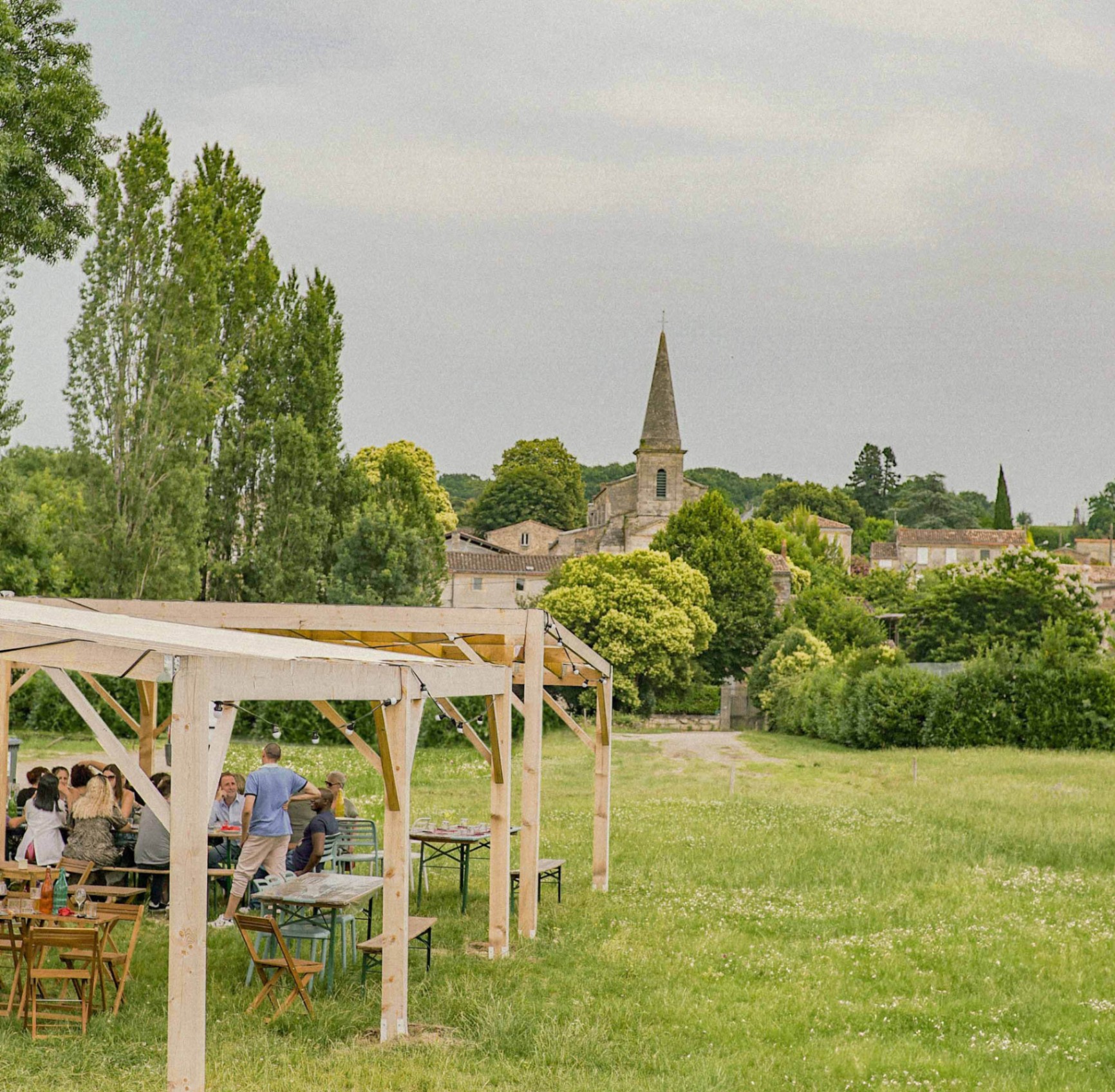 Vue panoramique de la Guinguette du Peyrat avec sa pergola en bois, ses convives et le clocher de Plassac en arrière-plan. - réalisé par Agence Kulte